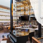 Elegant hotel lobby with bar, seating areas, and grand piano under a glass atrium.
