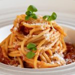 Spaghetti with tomato sauce, topped with grated cheese and fresh basil leaves in a white bowl.