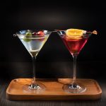 Two colorful martini cocktails on a wooden serving tray against a dark background.