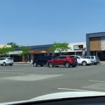 Strip mall with various storefronts and parked cars under a clear blue sky.