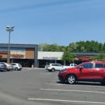 Strip mall with various storefronts and parked cars under a clear blue sky.