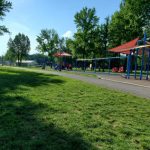 Sunny park scene with green grass, trees, and playground equipment under a blue sky.