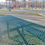 Green mesh fence in foreground overlooking a park playground and trees on a sunny day.