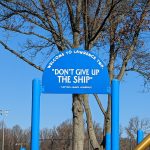 Blue sign reading "DON'T GIVE UP THE SHIP" under a large, bare tree against a blue sky.