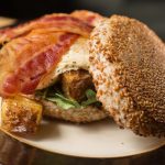 Gourmet burger with crispy bacon, sesame seed bun, and patty on a dark plate.