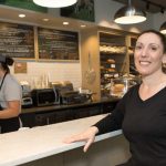 Woman smiling behind a counter in a bright, modern cafe setting.