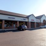 Strip mall featuring Investment Bank, Penlar Pharmacy, and other storefronts under a clear sky.