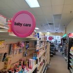Pink "baby care" sign in a brightly lit retail store aisle with shelves of baby products.