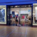 Retail store window display featuring denim and mannequins, with interior view of clothing racks.
