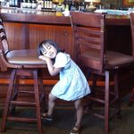 Young girl resting her head on a bar stool inside a dimly lit, upscale restaurant.