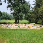Children play on a sandpit area surrounded by grass and trees in a park.