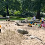 Children play with toy construction vehicles on a sandy area surrounded by trees.