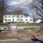 Historic white farmhouse with green shutters, framed by large trees on a sunny day.
