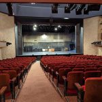 Theater auditorium with rows of red seats facing a large, dark stage.