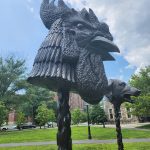 Dark, textured sculpture of a dragon head and smaller creature on a grassy campus.