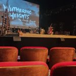 Concert stage with large screen displaying "Nuttering Heights by Emily Brout" and red audience seats.