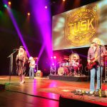 Band performs live on stage under bright lights with "Tusk" Fleetwood Mac tribute banner.