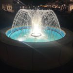 Nighttime fountain spraying water brightly in a circular basin with surrounding architecture.