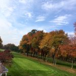 Autumn trees line a manicured green golf course under a bright blue sky.
