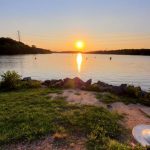 Sunset over calm water with a silhouetted shoreline and grassy foreground.