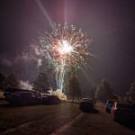 Fireworks explode over a dark, grassy field with trees lining the horizon at night.