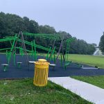 Green playground equipment set in a grassy park area with trees in the background.