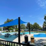 Blue shade sail over a sunny outdoor splash pad with surrounding greenery.