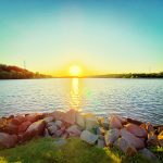 Sunset over a calm lake with rocks lining the shore under a bright blue sky.