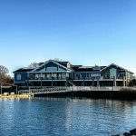 Blue waterfront house complex with decks overlooking a calm lake under a clear sky.