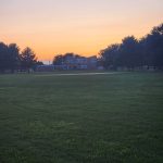 Sunset over a green field with a house and trees silhouetted against the orange sky.