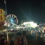 Nighttime carnival scene with a Ferris wheel, food stalls, and crowds under bright lights.
