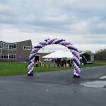 Purple and white balloon archway entrance to an outdoor event with a building in the background.