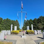 War memorial with flagpole and flags surrounded by rows of white headstones under a blue sky.