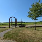 Outdoor park scene with a metal archway, picnic tables, and green fields under a blue sky.