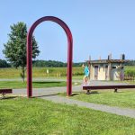 Red archway frames a park scene with grass, trees, and a small building under a blue sky.