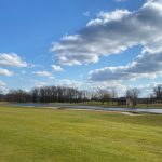Expansive green golf course with a pond under a blue sky dotted with white clouds.