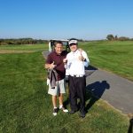 Two smiling men in golf attire give thumbs up on a sunny golf course.