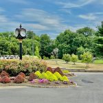 Park entrance with a decorative clock, lush green trees, and flowering bushes under a blue sky.