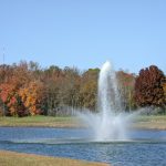 Water fountain spraying high from a pond surrounded by autumn trees under a blue sky.