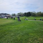 Golfers on a green course with trees and buildings in the background under a cloudy sky.