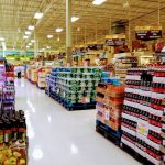 Grocery store aisle with refrigerated drinks, stacked produce, and shelves of packaged goods.