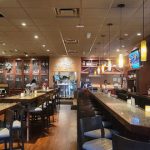 Interior view of a modern bar and restaurant with wooden tables, seating, and glowing liquor displays.