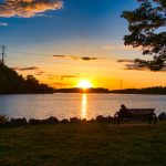 Sunset over a calm lake with silhouetted trees and a park bench.