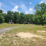 Park scene with grassy field, winding path, playground structure, and trees under a blue sky.