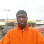 Man in orange jacket and black cap standing outdoors near a commercial building.