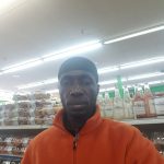 Black man in orange jacket stands in a brightly lit grocery store aisle.