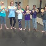 Group of young women posing indoors, holding small cards at a martial arts training center.