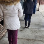 People walking down a covered walkway outside a storefront with brick walls.
