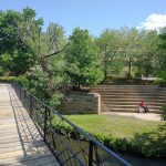 Wooden boardwalk overlooks a stream with tiered stone steps and green trees in the background.