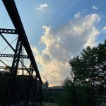 Steel bridge structure against a dramatic, cloudy blue sky with trees in the foreground.